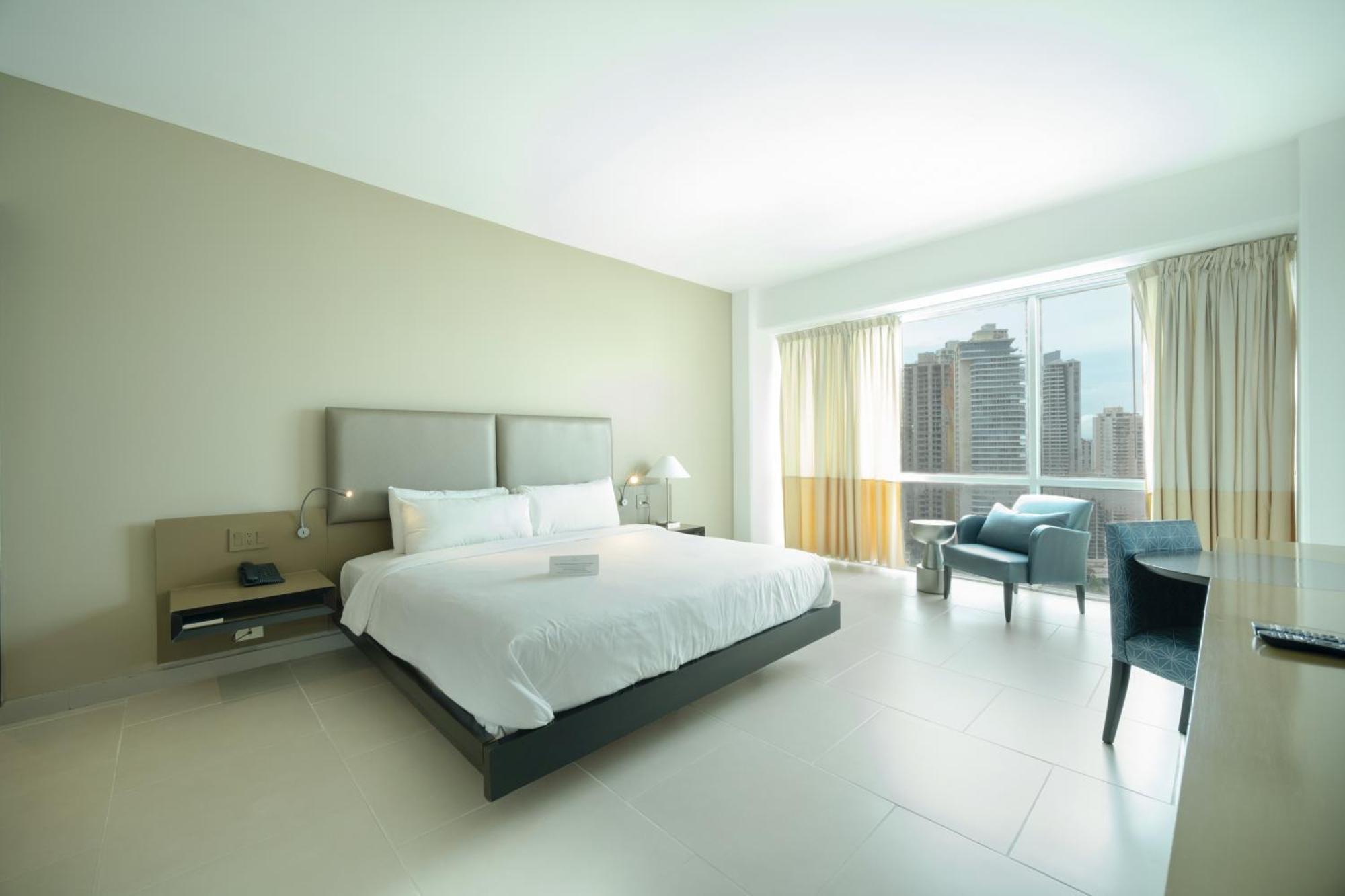 Decapolis Hotel פנמה סיטי מראה חיצוני תמונה The photo shows a modern hotel room with a minimalist design. There is a large bed with a light-colored duvet and two decorative pillows. Next to the bed, there is a nightstand with a lamp. On the opposite side, a comfortable chair is placed near a l