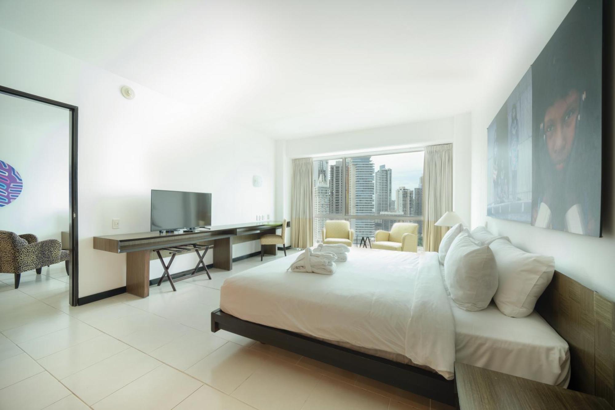 Decapolis Hotel פנמה סיטי מראה חיצוני תמונה The photo depicts a modern hotel room featuring a large bed with white linens and several pillows. There is a television mounted on the wall opposite the bed, and a desk with a chair beside it. Large windows provide a view of a city skyline, with two