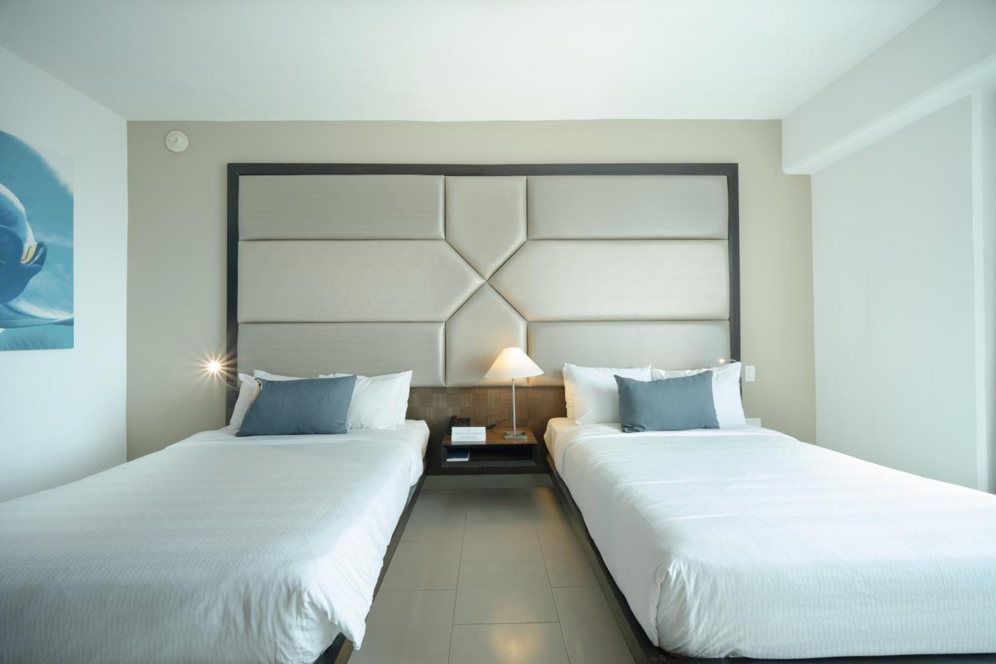 Decapolis Hotel פנמה סיטי מראה חיצוני תמונה The photo shows a modern hotel room with two neatly made beds, each with white bedding. The beds are positioned next to each other, with a contemporary headboard behind them featuring a geometric design. There is a small bedside table between the bed