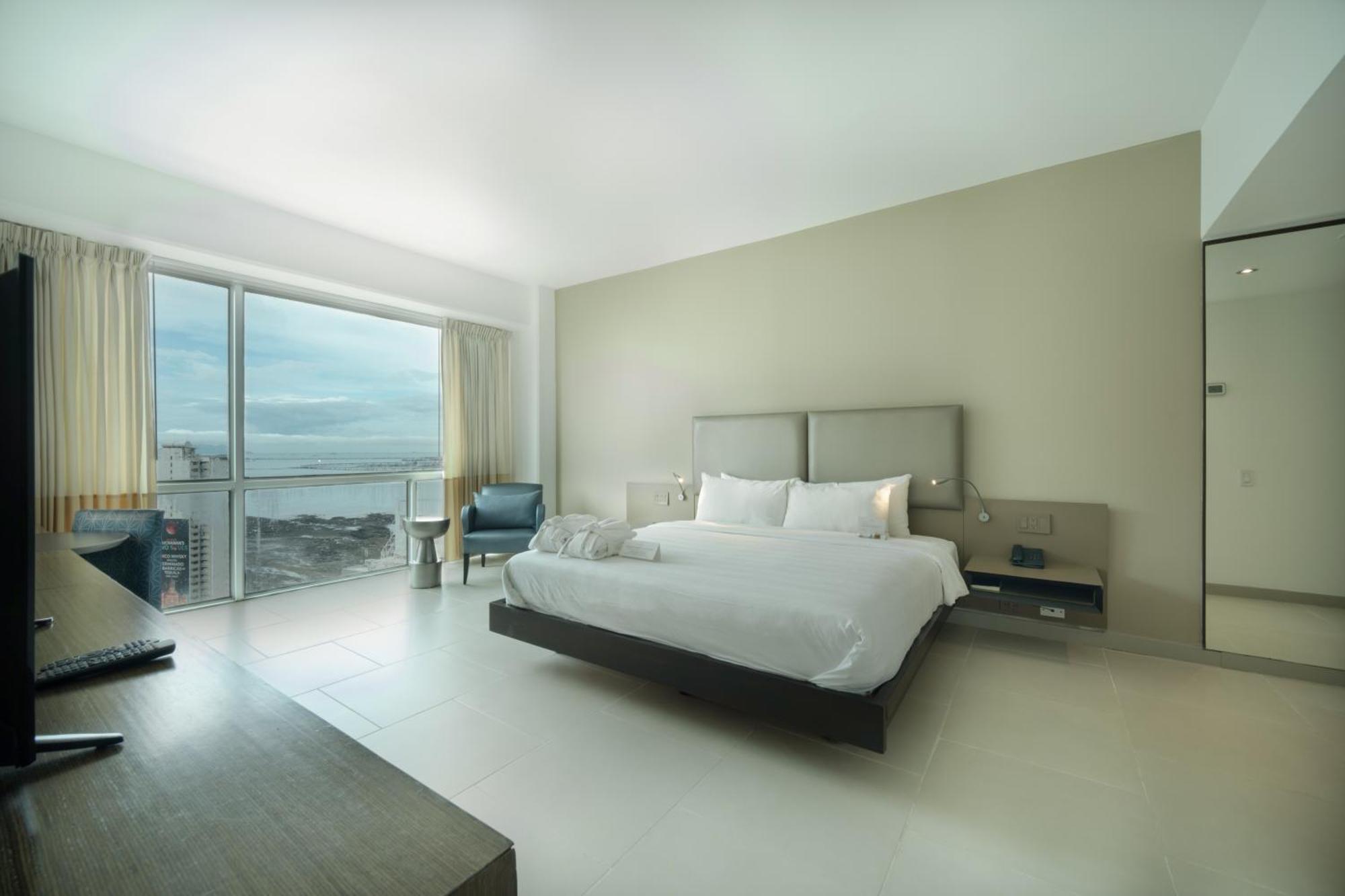 Decapolis Hotel פנמה סיטי מראה חיצוני תמונה The photo shows a modern and minimalist hotel room. It features a large bed with white bedding, complemented by neutral-colored walls. There is a sleek wooden desk and a chair in the corner, along with a small seating area by a window that looks out 