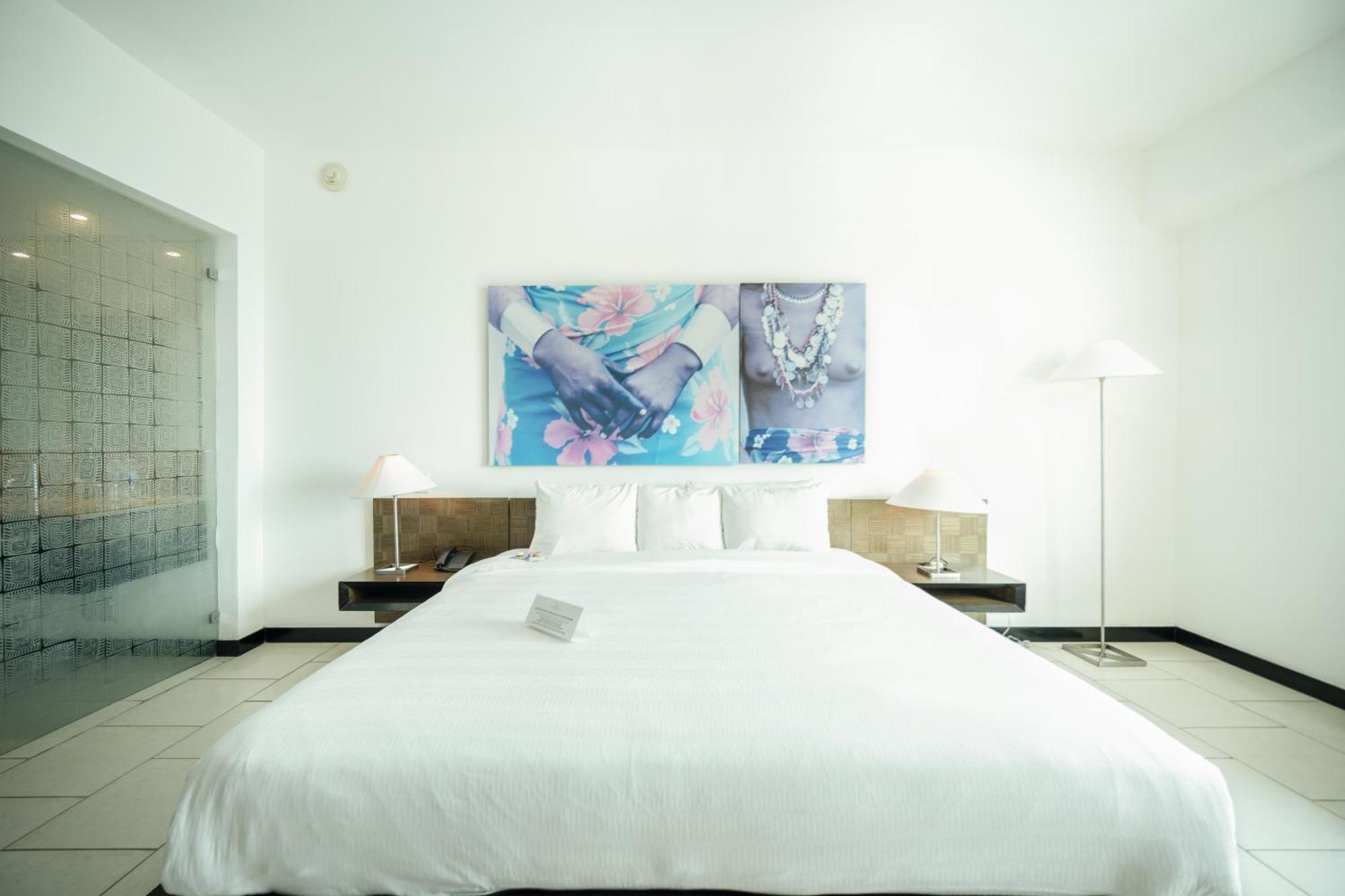 Decapolis Hotel פנמה סיטי מראה חיצוני תמונה The photo shows a modern bedroom with a clean and minimalist design. The bed is large with a white comforter and several pillows. On the wall above the bed, there are two framed art pieces featuring floral and jewelry motifs, adding a touch of color 
