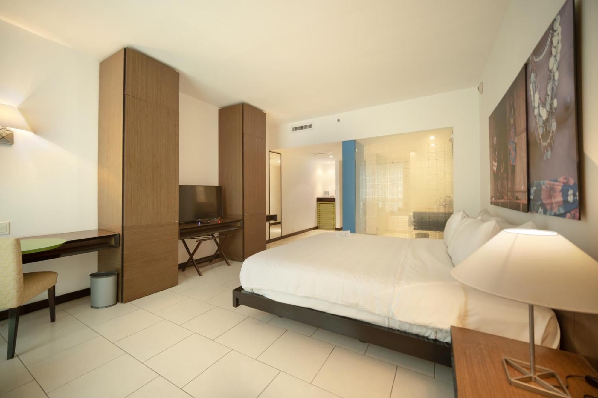 Decapolis Hotel פנמה סיטי מראה חיצוני תמונה The photo shows a modern hotel room. It features a large bed with white bedding, a wooden headboard, and bedside tables with lamps. There is a wooden wardrobe or storage unit on one side, along with a desk and a chair. A flat-screen TV is mounted on 