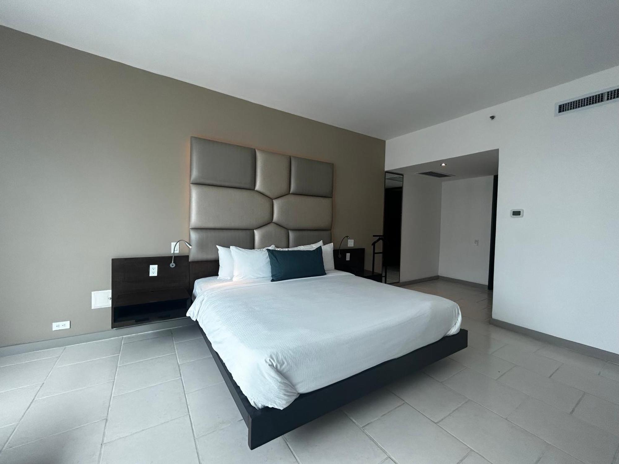Decapolis Hotel פנמה סיטי מראה חיצוני תמונה The photo shows a modern hotel room featuring a spacious, minimalist design. In the center, there is a large bed with a white duvet and pillows, positioned against a light gray accent wall that has a padded headboard. To the side, there is a simple n