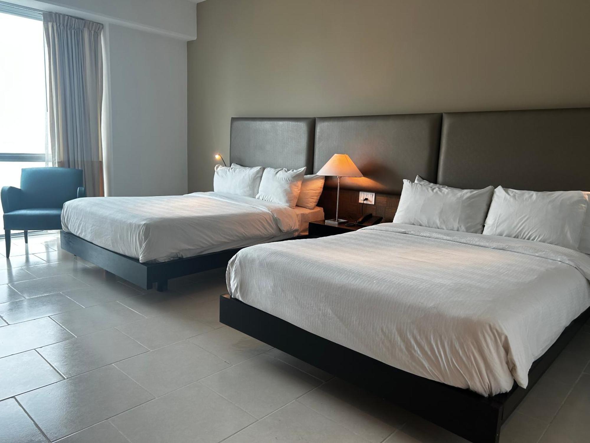 Decapolis Hotel פנמה סיטי מראה חיצוני תמונה The photo shows a hotel room featuring two large beds with white beddings. There is a bedside table with a lamp next to one of the beds. A cozy chair is placed in the corner of the room. Large windows allow natural light to fill the space, and the wa