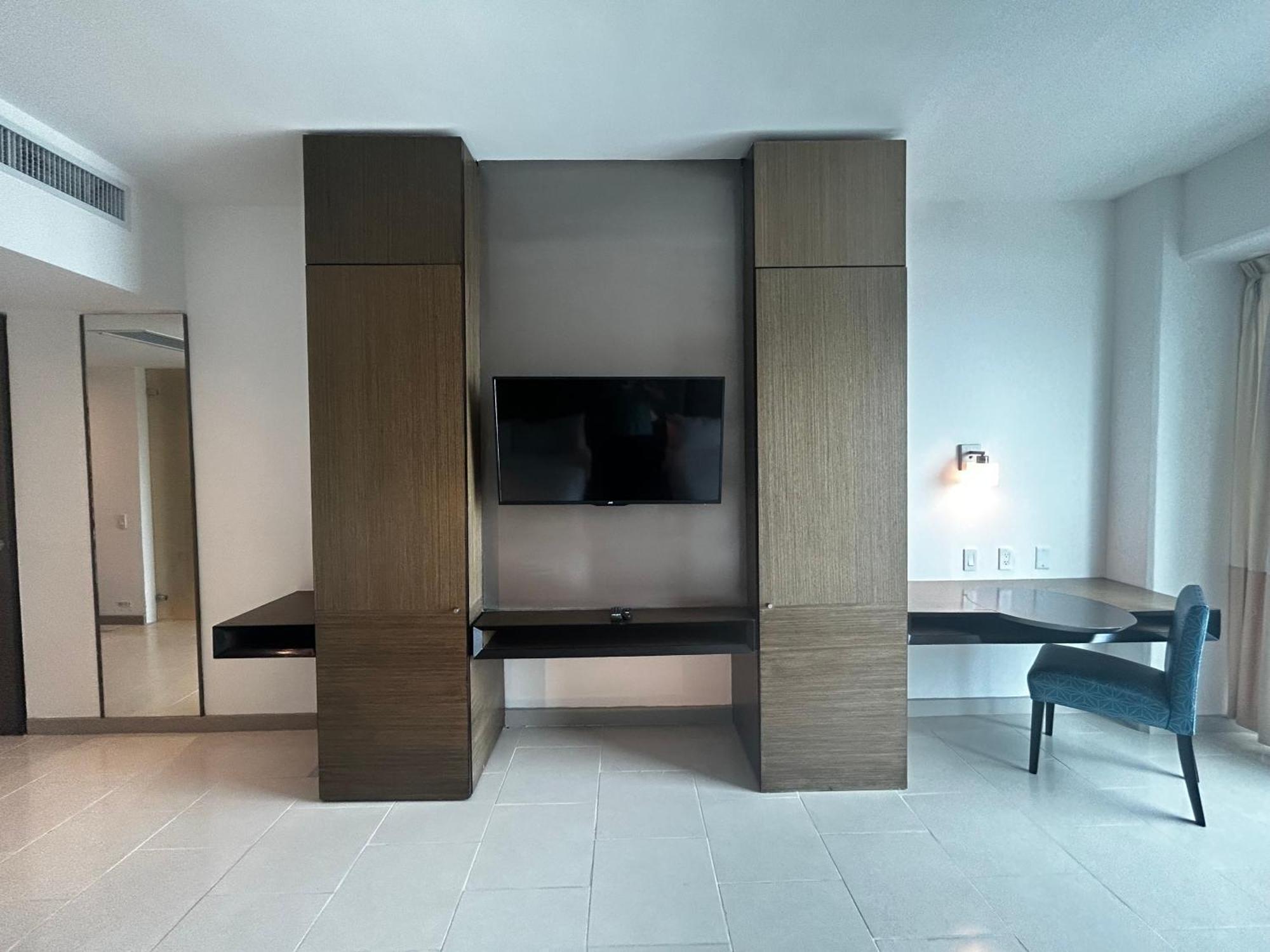 Decapolis Hotel פנמה סיטי מראה חיצוני תמונה The photo shows a modern interior space, likely a hotel or apartment room. Central to the image is a mounted flat-screen television on a wall, flanked by tall, narrow cabinets or panels made of a wood-like material. Below the TV, there is a long, dar