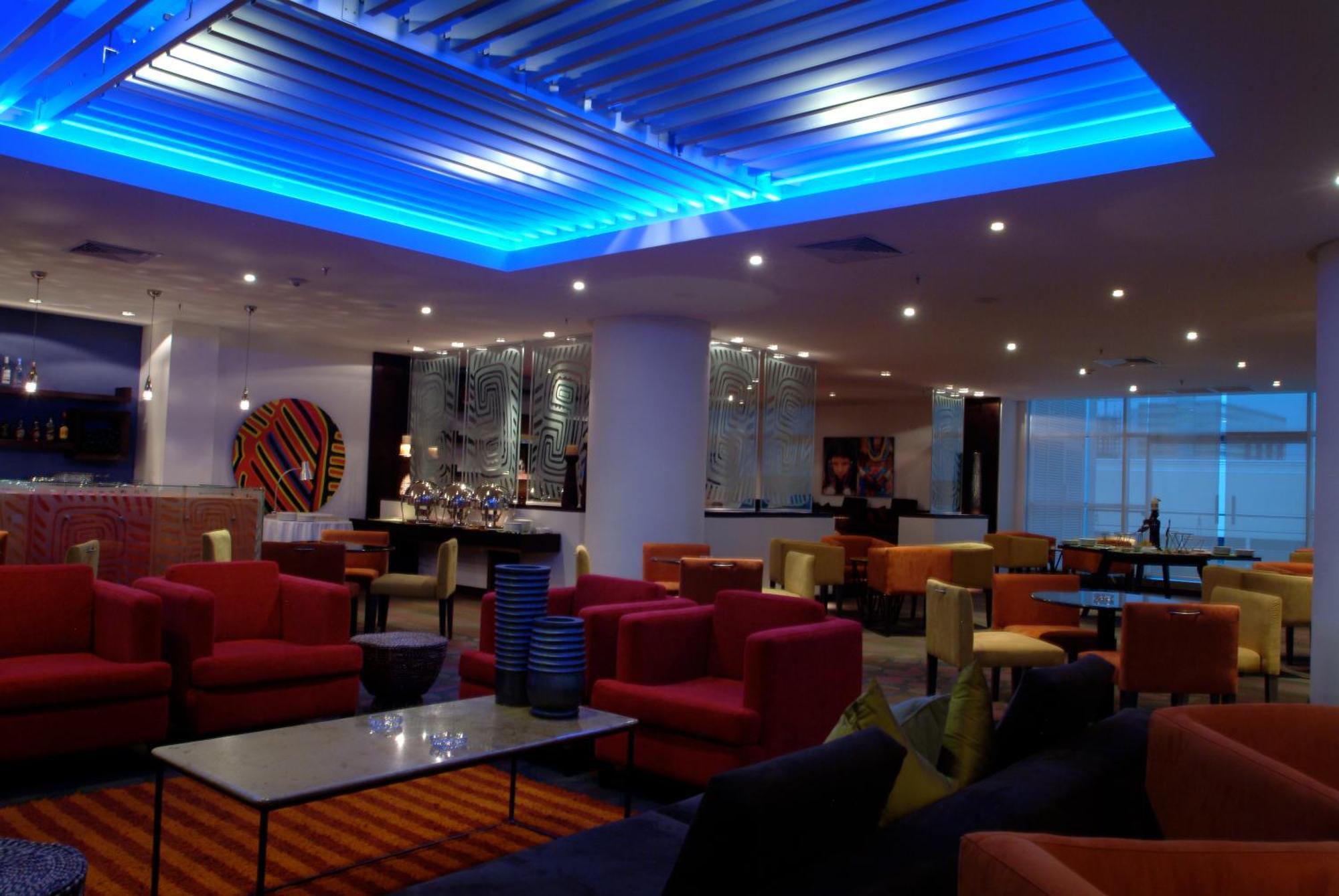 Decapolis Hotel פנמה סיטי מסעדה תמונה The photo shows a modern lounge or lobby area with various seating arrangements. The furniture includes colorful chairs and sofas, predominantly in shades of red, pink, and yellow, arranged around glass-topped tables. The ceiling features a blue illu